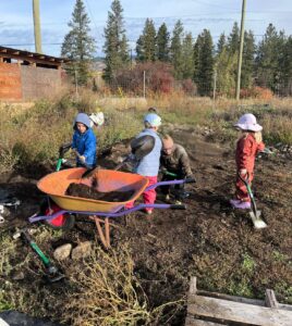 Four young children in jackets and rain gear shovel soil into an orange wheelbarrow in the school garden on an autumn day.
